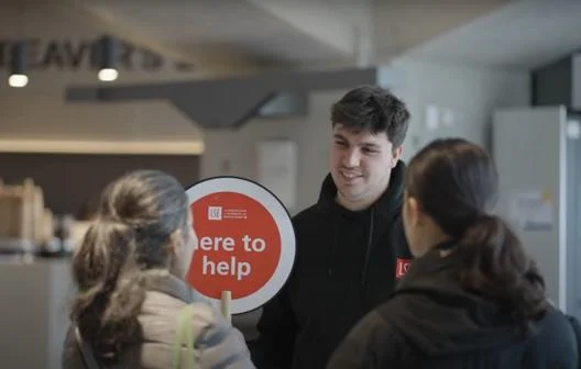 A Student Ambassador holding a 'here to help' sign talking to visitors.