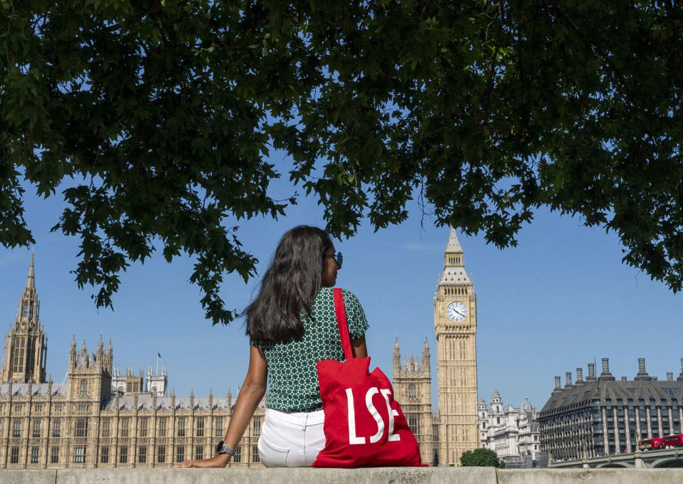 An LSE student with a red LSE tote bag sits by the river Thames overlooking Westminster on a sunny day
