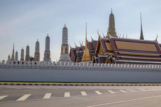 Temple complex with ornate spires and a white wall, viewed from across an empty street under a clear sky.