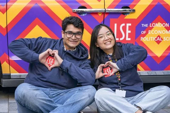 Two LSE students pose in front of a colourful LSE-branded vehicle, smiling and making heart shapes with their hands.