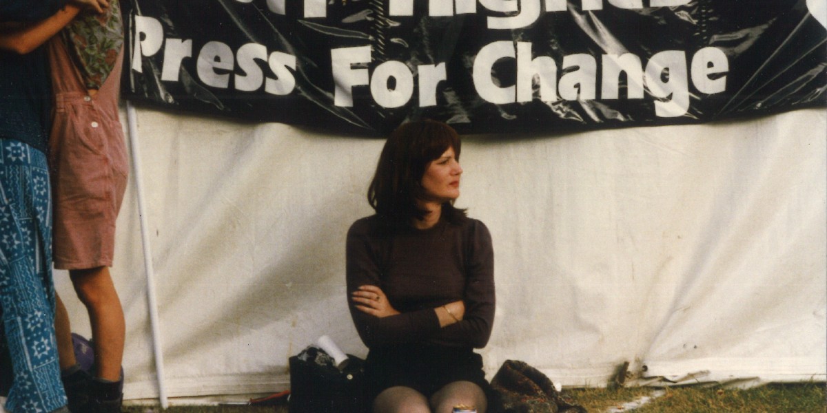 A person sat up against a tent with a sign behind them. Sign reads "Press For Change".