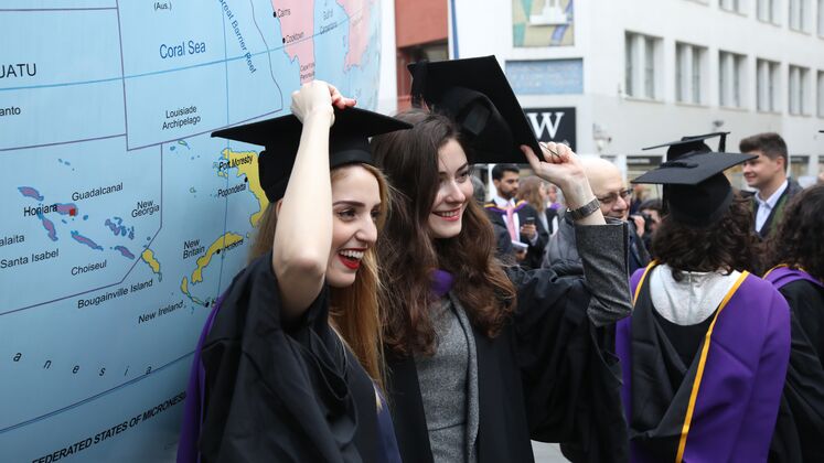 Two women students get a photo taken of themselves in their graduation gowns and caps beside Mark Wallinger's 'World Turned Upside Down'