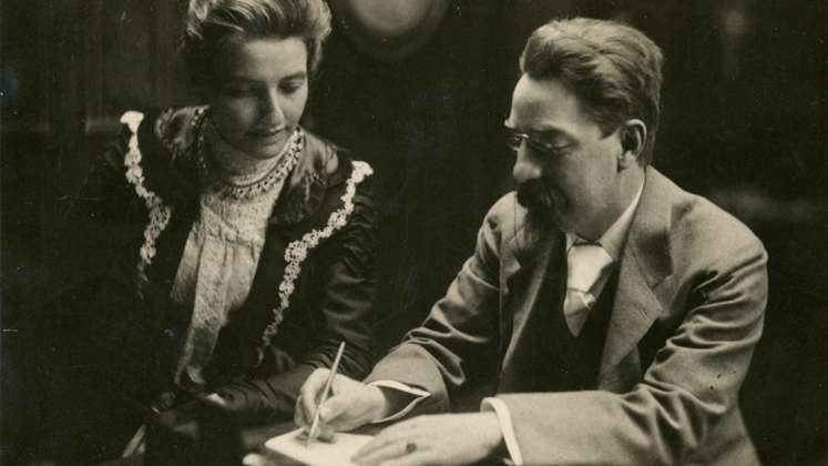 Sidney Webb writes in a book as Beatrice looks over his shoulder