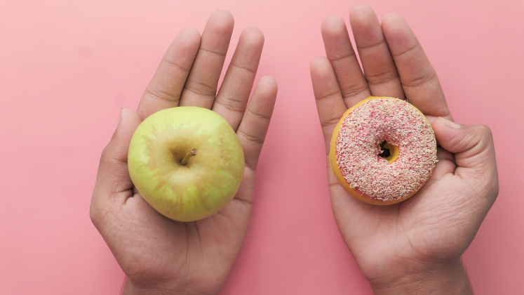 Two hands on pink background one holding an apple and the other a doughnut_sourced via Canva 747x420