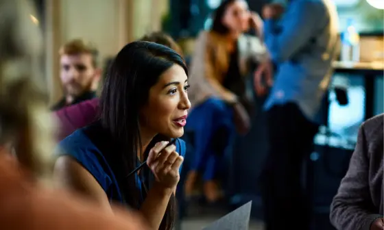 Woman in a busy office with a notebook in hand speaking to someone out of shot
