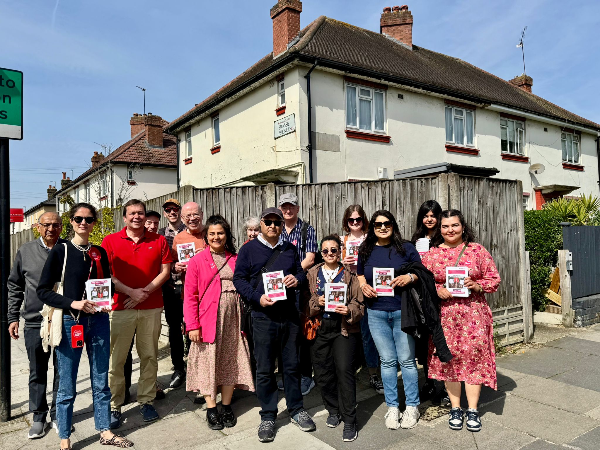 Group standing on a sunny street in front of houses and a wooden fence, holding campaign leaflets.