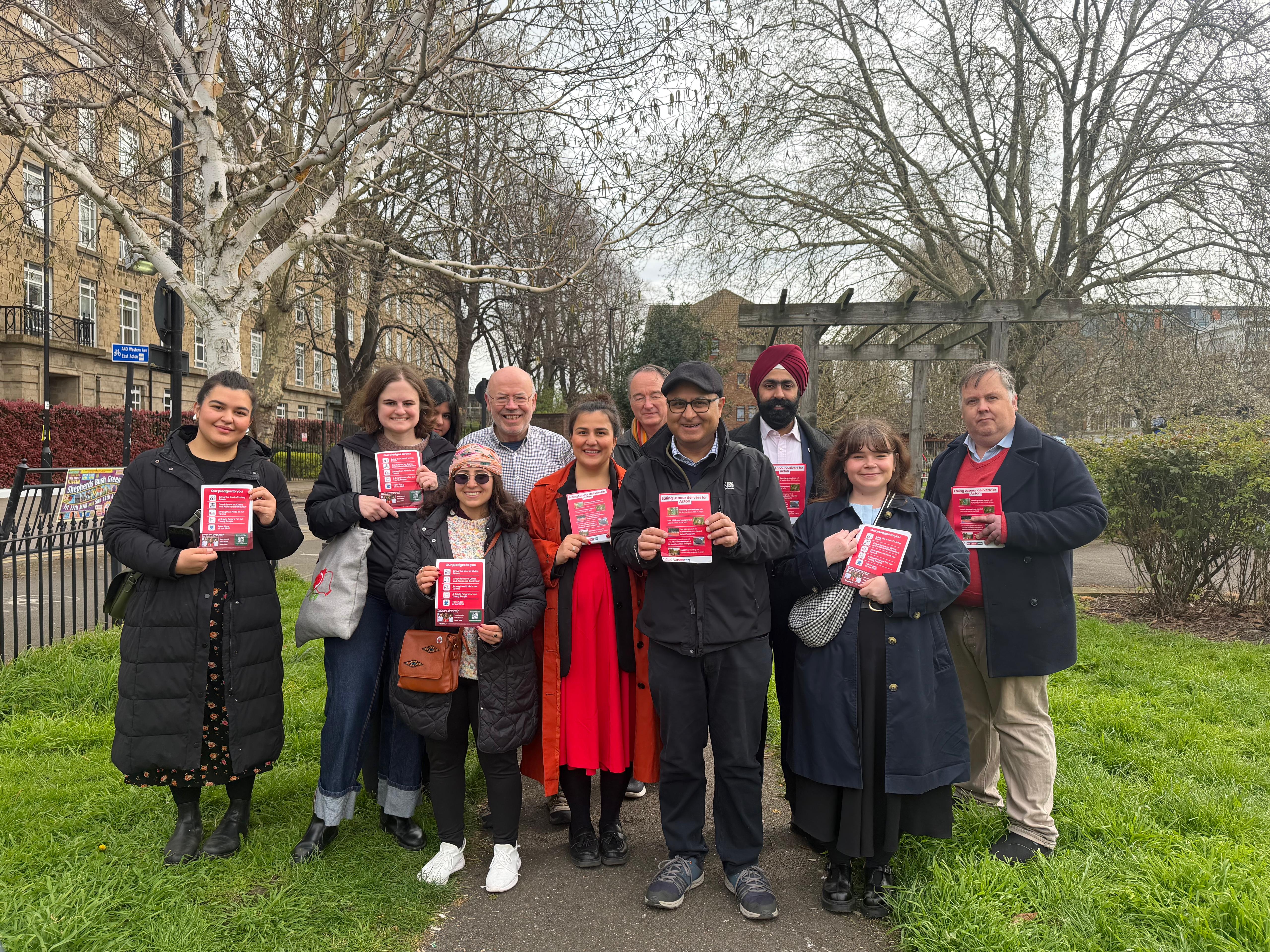 Group standing in a park holding red campaign leaflets, with trees and buildings behind them.