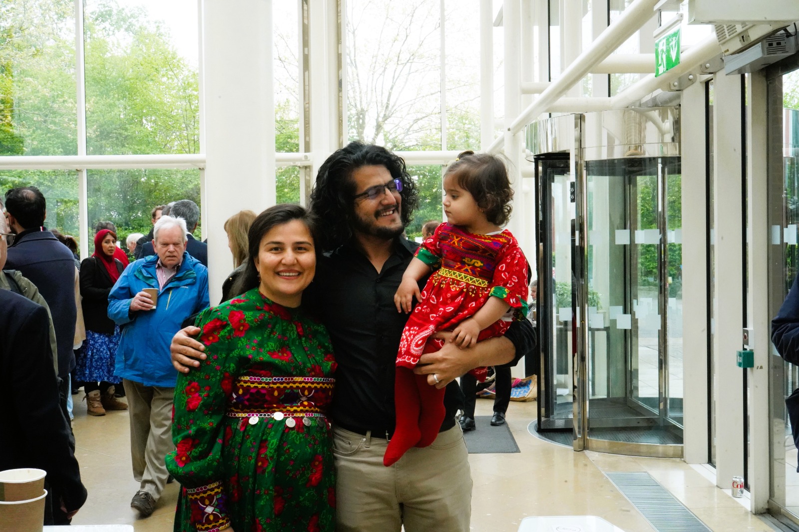 Two adults and a child standing inside a bright building near a glass entrance, with people gathered in the background.