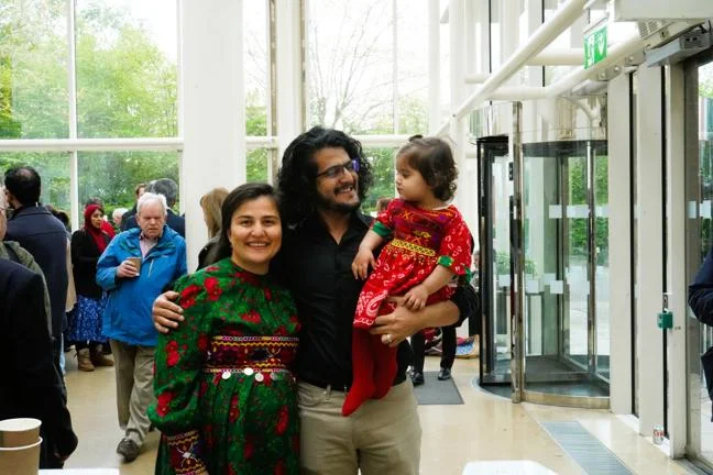 Two adults and a child standing inside a bright building near a glass entrance, with people gathered in the background.