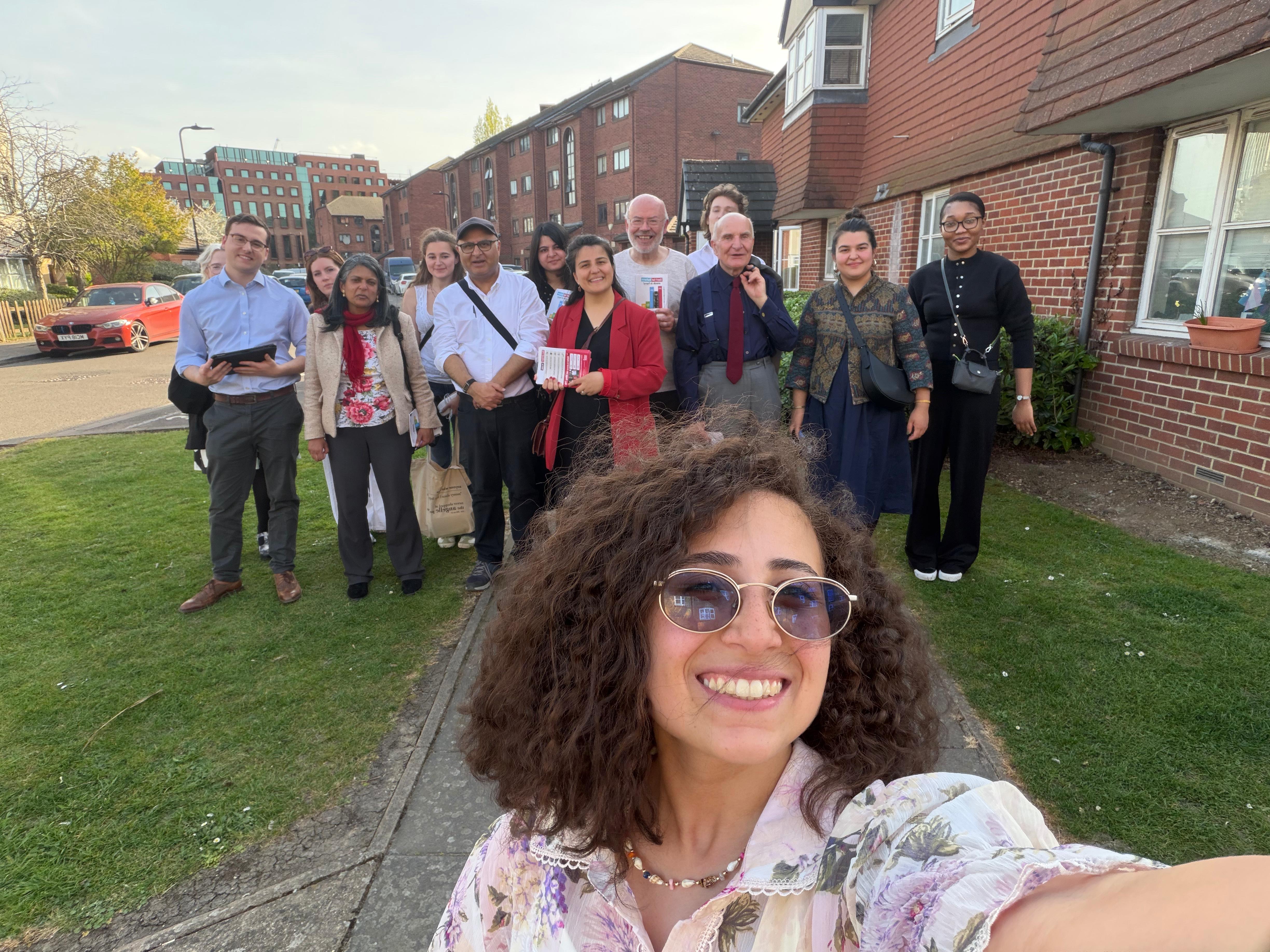 Person taking a selfie in the foreground with a group standing on a residential street behind them, some holding campaign materials.