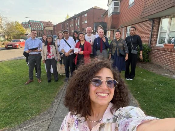 Person taking a selfie in the foreground with a group standing on a residential street behind them, some holding campaign materials.