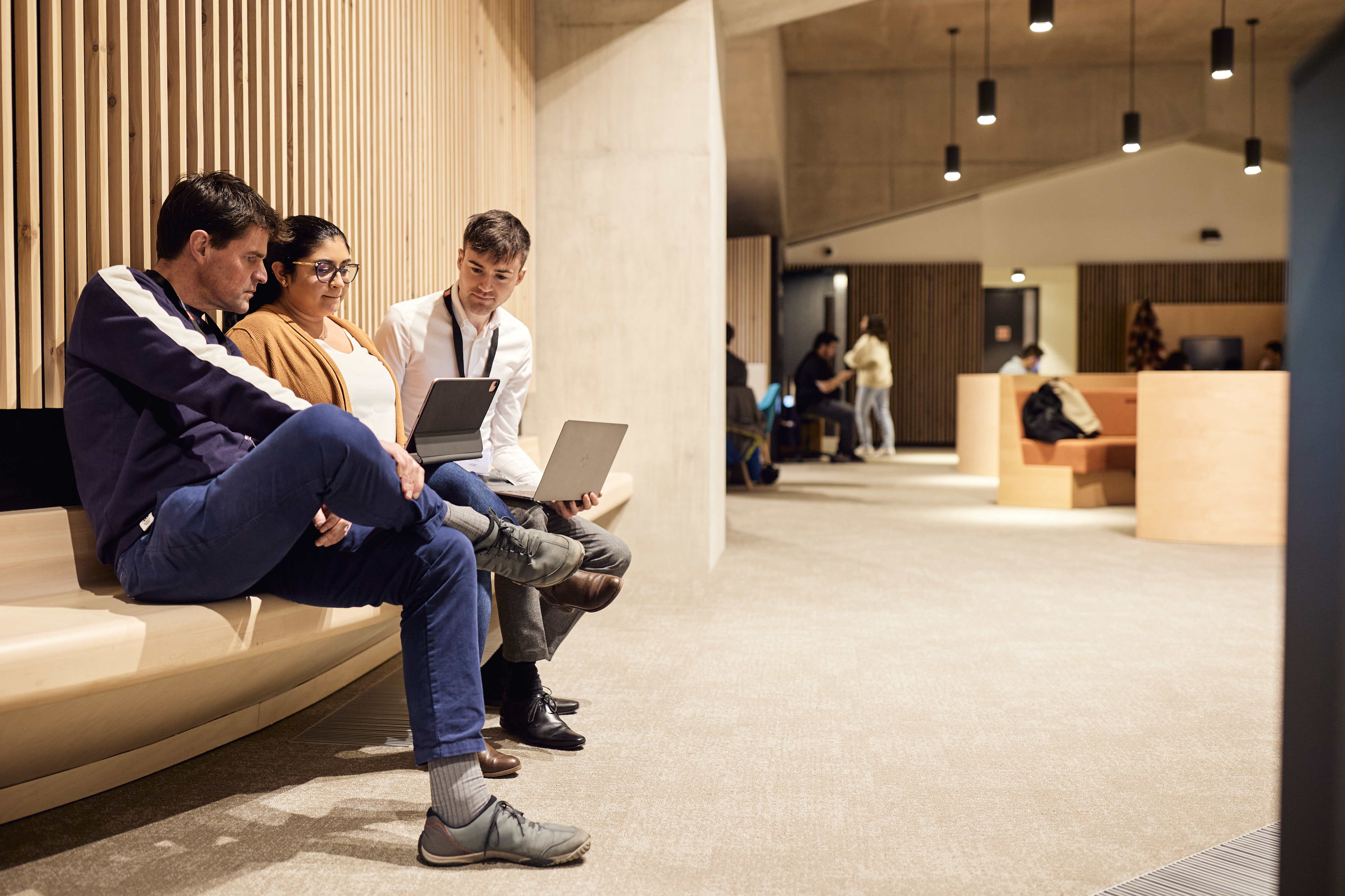 Three students collaborating on laptops while seated on a bench outside lecture theatre.