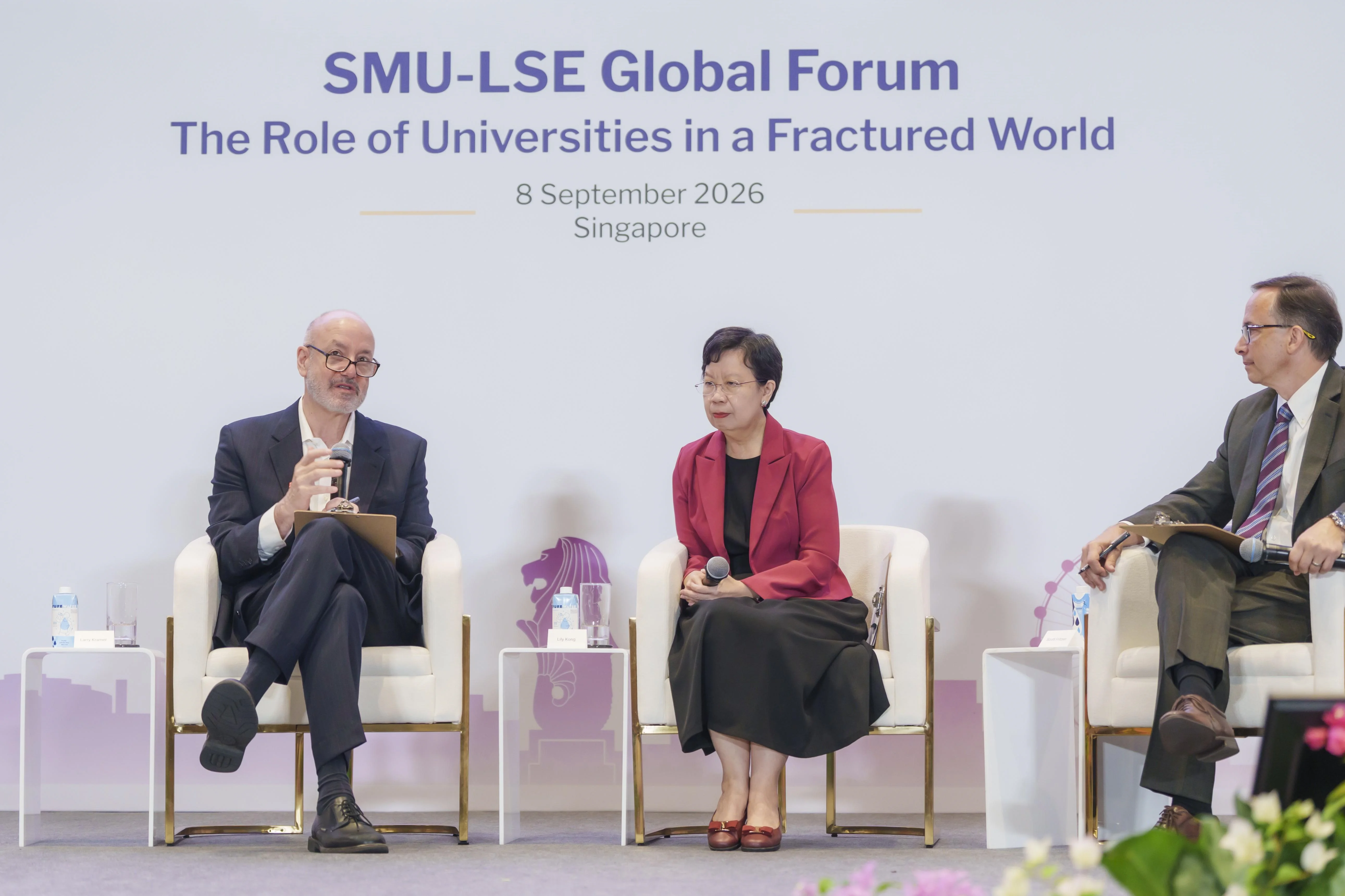 LSE President Larry Kramer sits on a chair on a stage, holding a microphone. He is joined by SMU President Lily Kong and SMU Vice President Scott Fritzen, who are also sitting in chairs on the stage, facing Kramer.