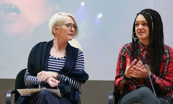 Two speakers at an event in sit down in front of a screen
