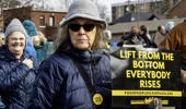Protesters marching, with a woman holding a sign reading “Lift from the bottom, everybody rises.”