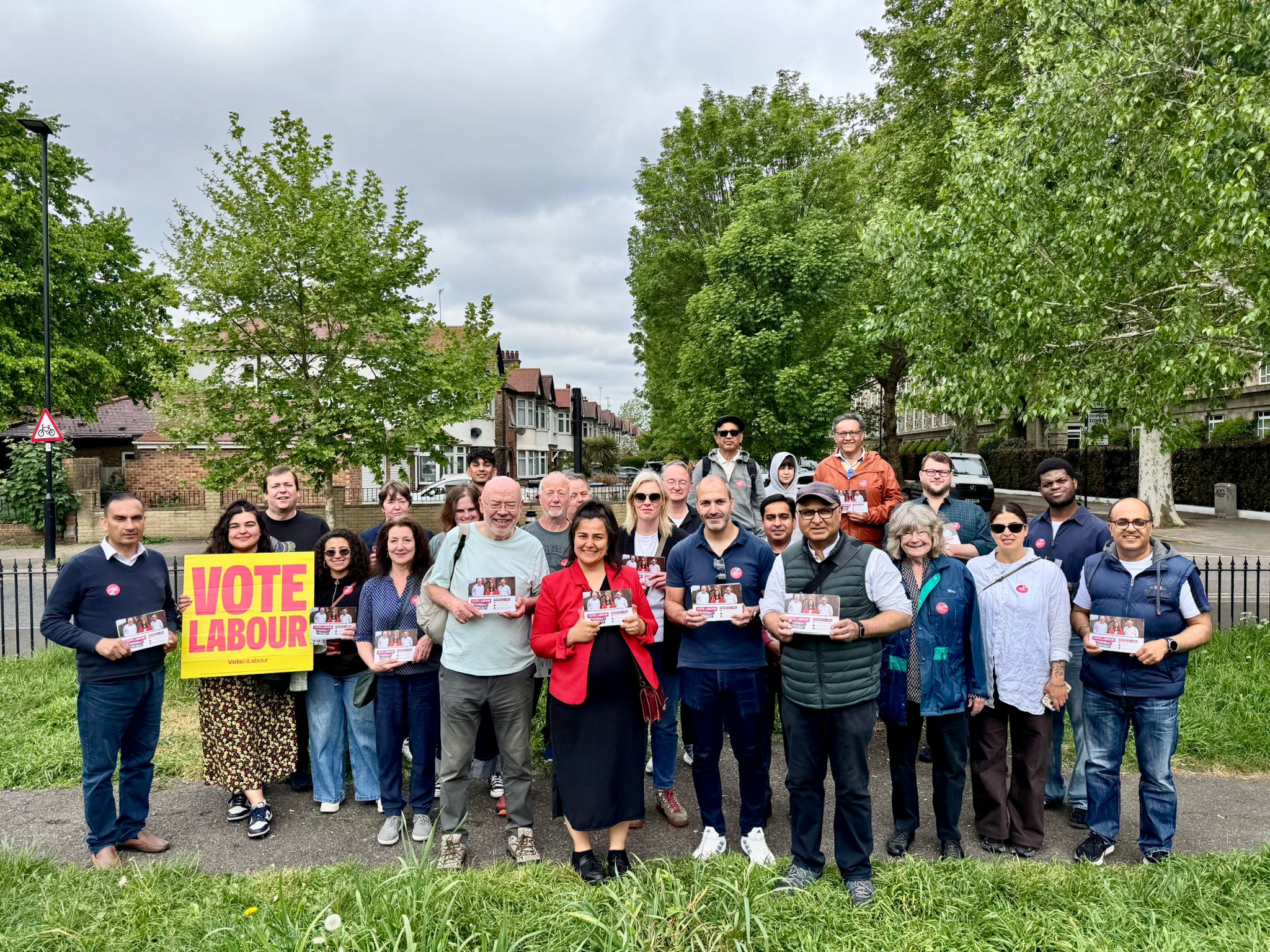Group of people on a residential street holding campaign leaflets and a yellow “Vote Labour” sign, with trees and houses in the background.