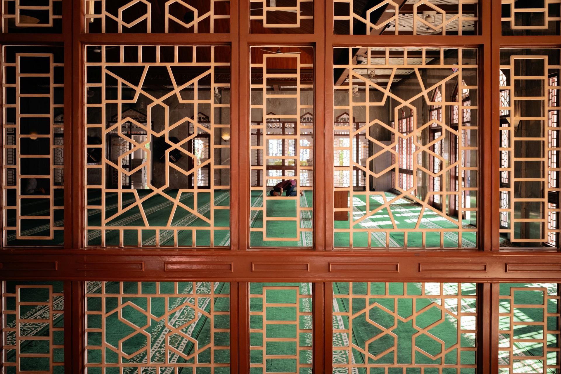 Intricate wooden screens frame a mosques interior, where a man is praying