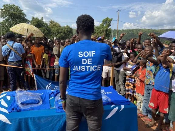 A group of people are gathered behind a desk. In the foreground a man wears a blue t-shirt reading "Soif de justice".