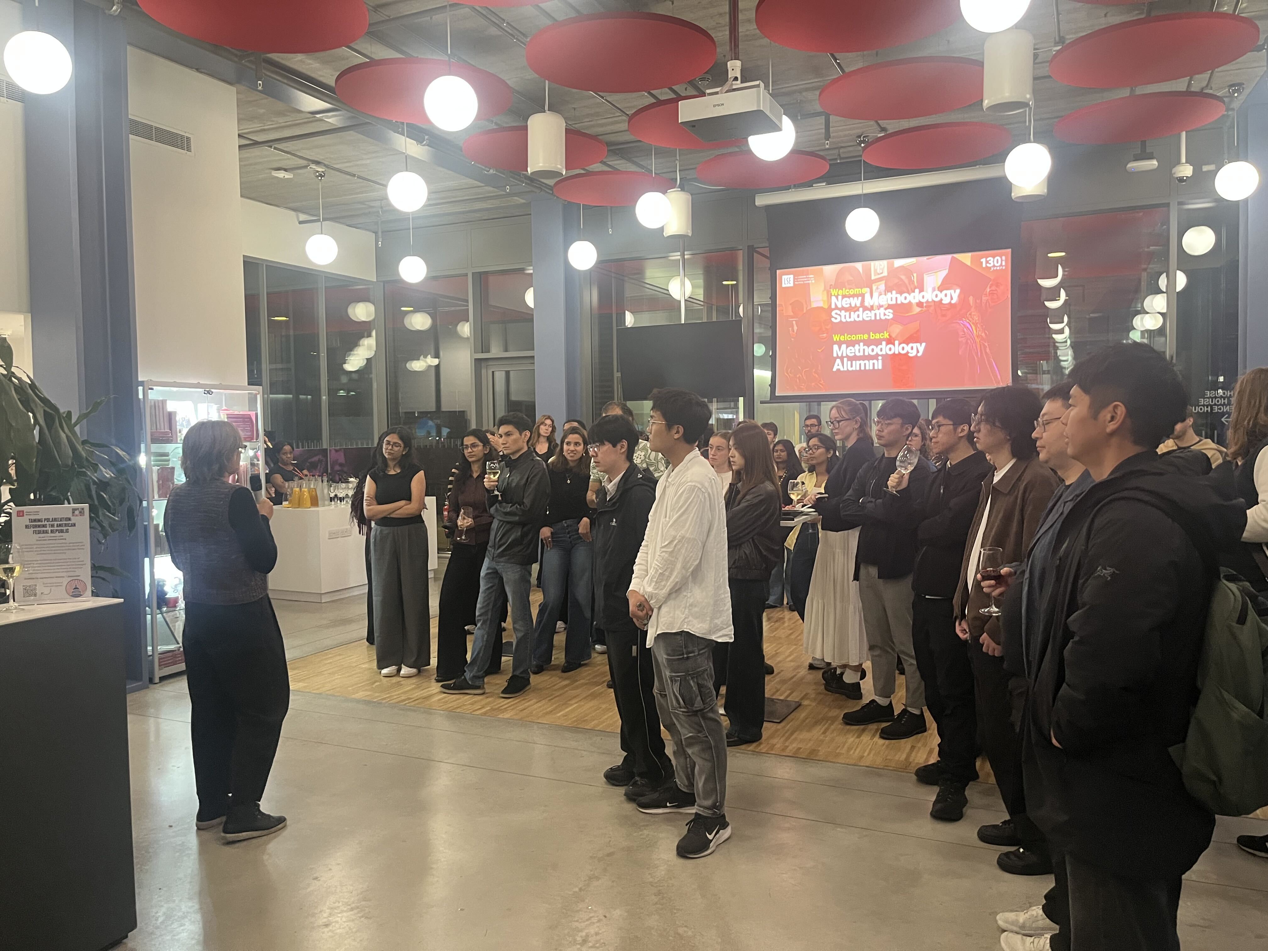 A group of people dressed formally stands in a well-lit event space, listening to a speaker at the front.