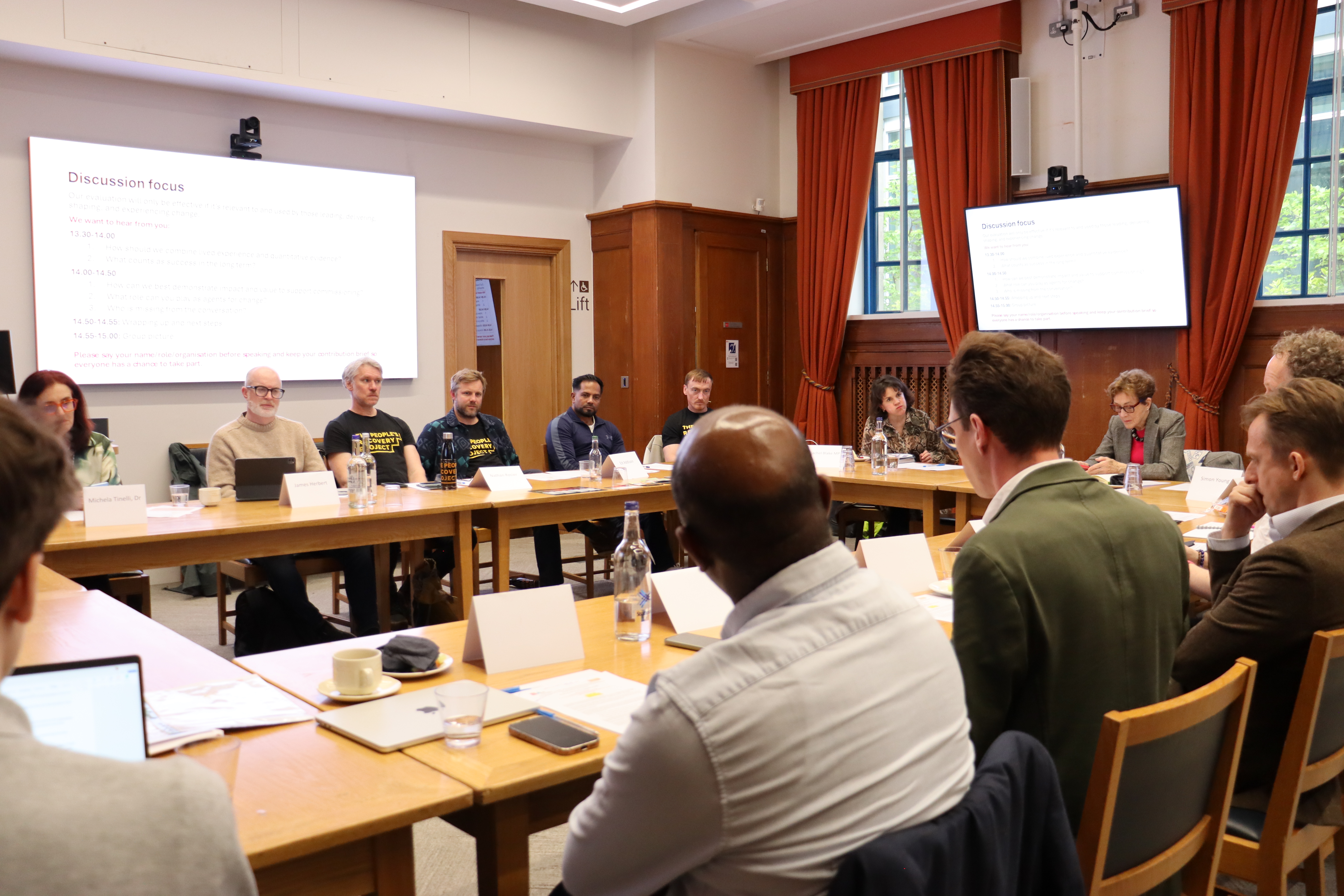 Attendees seated around tables in a meeting room, taking part in the group discussion. Khosru is sat at the top table, with other TPRP community and team members. Papers, laptops and drinks are on the tables, and a presentation slide is shown on a large screen at the front.