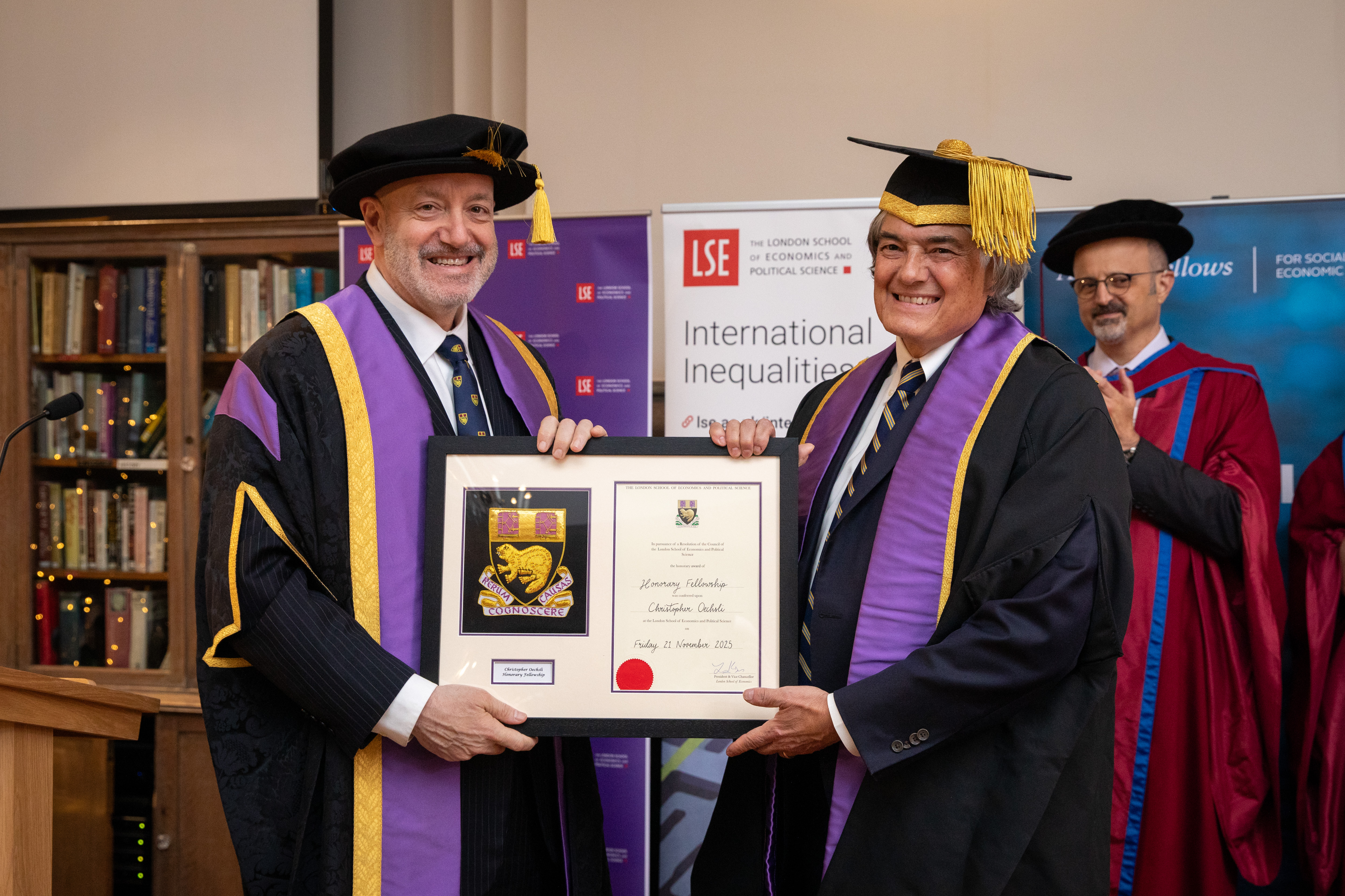 Professor Larry Kramer presents Chris Oechsli with an Honorary Fellowship. Both are wearing caps and gowns. 
