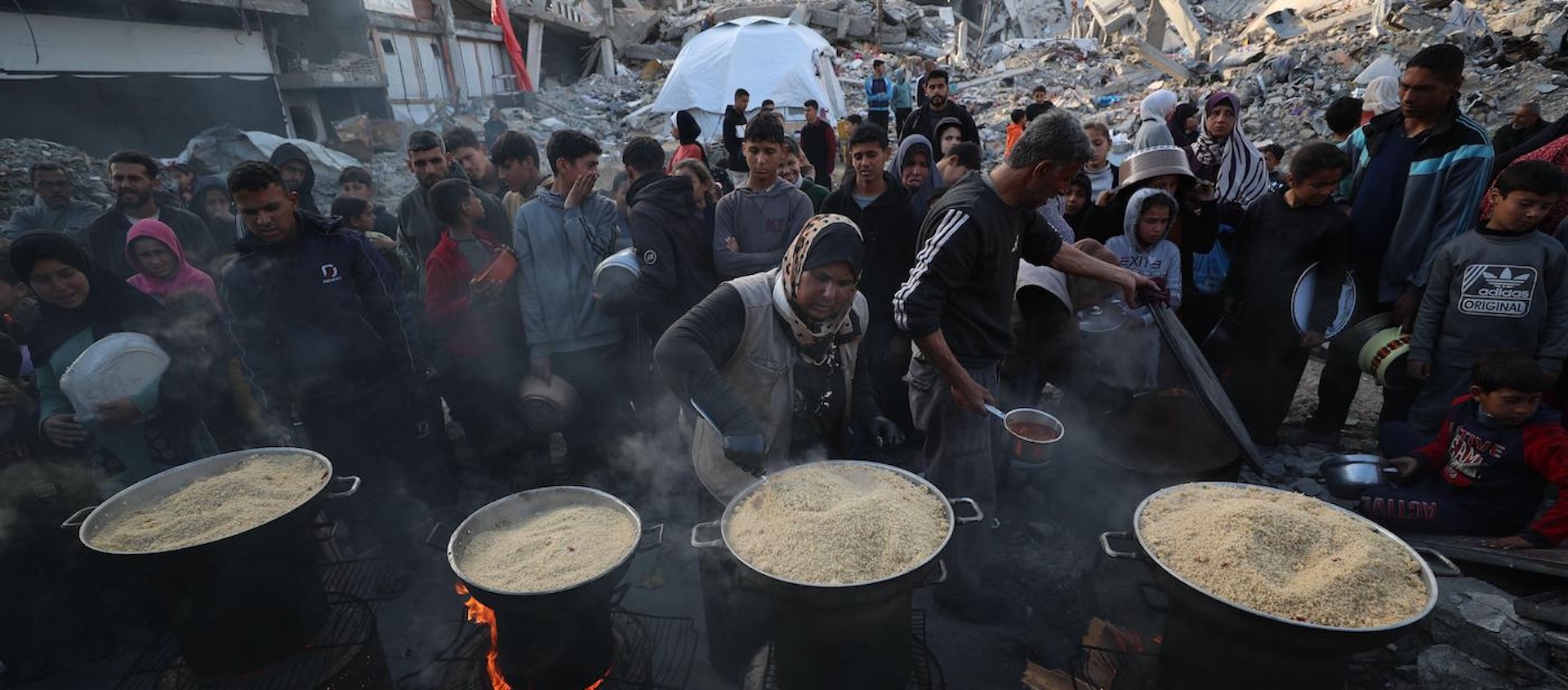 Women serving food from four large pots to a group of people, among the rubble of destroyed buildings in Gaza.  