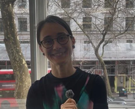 Kinga stands and smiles at the camera, holding a mini lapel microphone