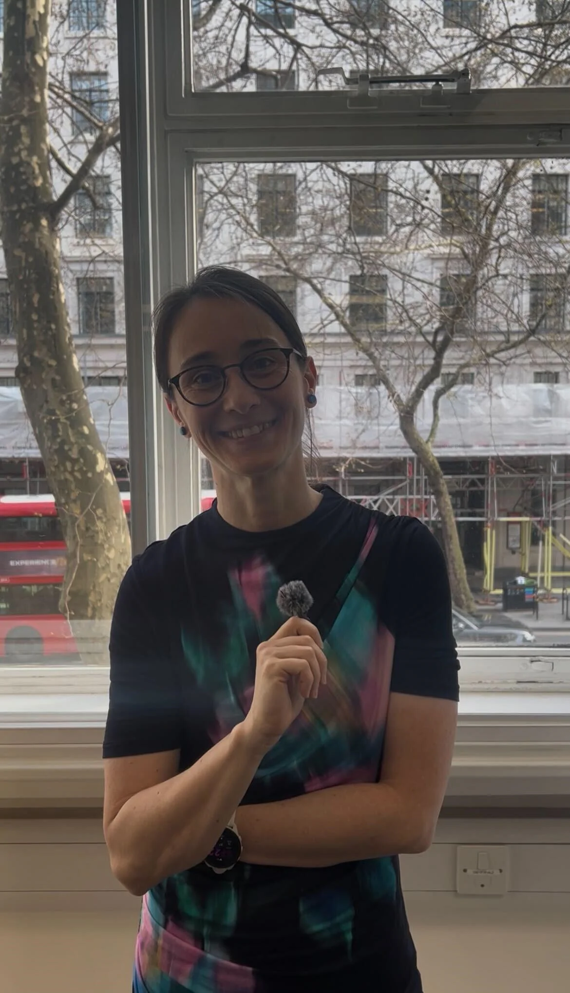 Kinga stands and smiles at the camera, holding a mini lapel microphone