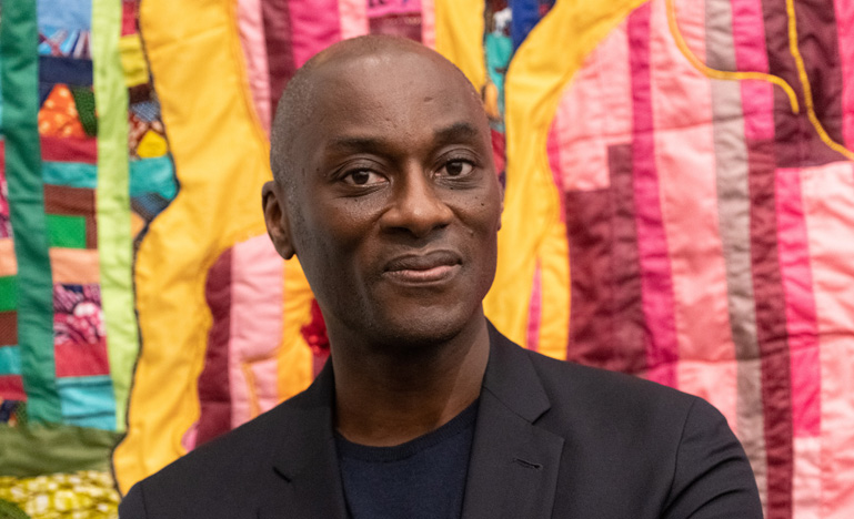 Profile image of Ekow Eshun standing in front of colourful textiles