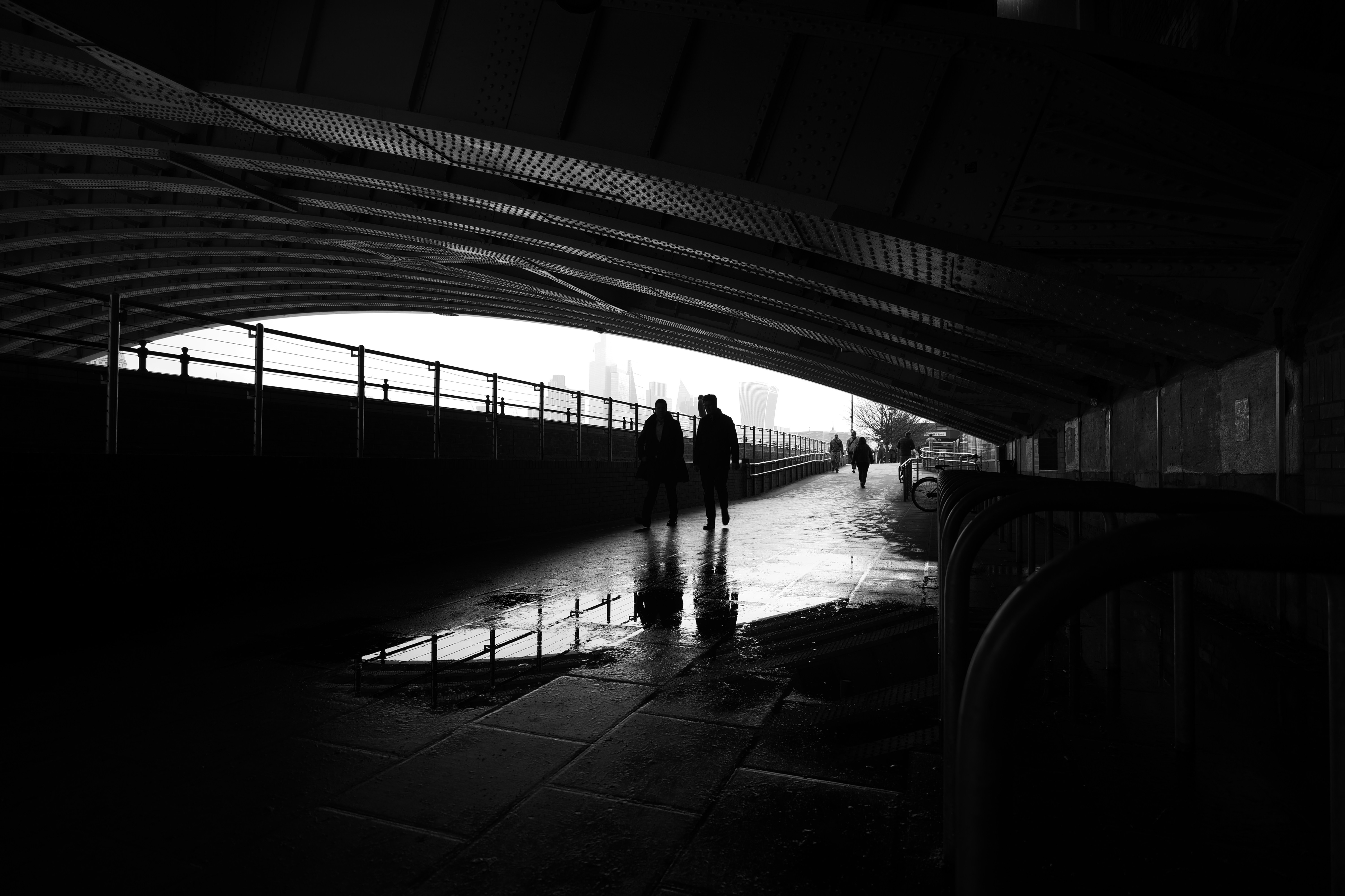Black-and-white photograph of a covered walkway beneath a large metal bridge. Two people are walking side by side in silhouette along a wet pavement, with reflections visible in puddles. More people are visible further ahead near the open end of the walkway, where daylight is much brighter and the London skyline is visible. 