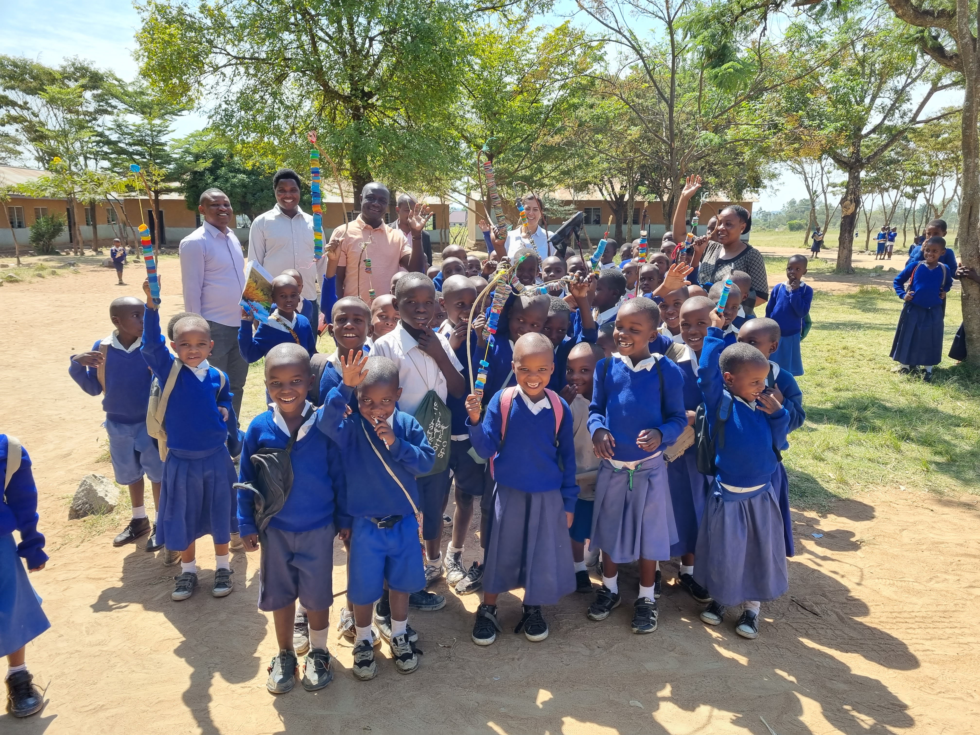 a group of children stand and smile at the camera, some adults in the background stand and wave too