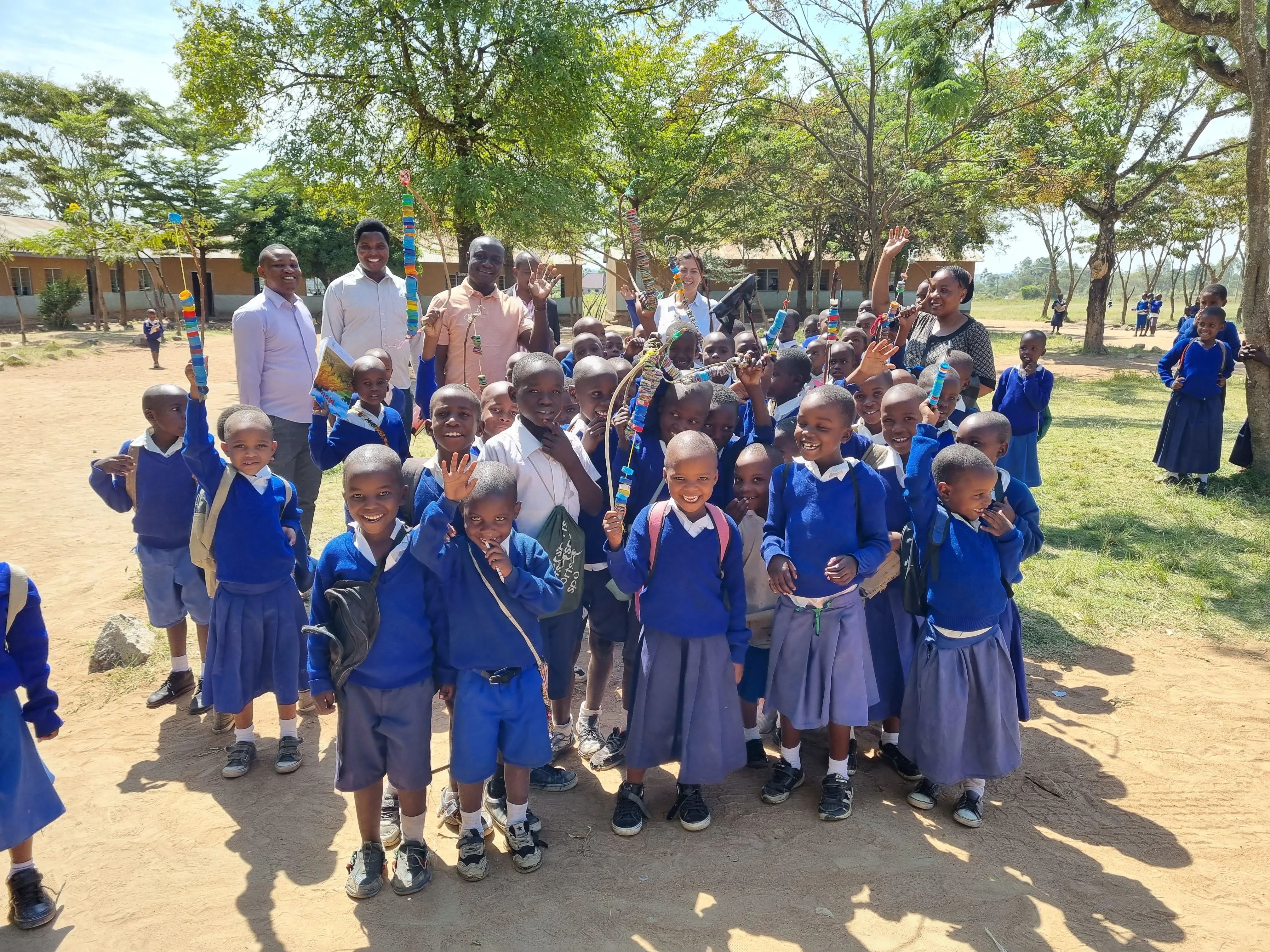 a group of children stand and smile at the camera, some adults in the background stand and wave too