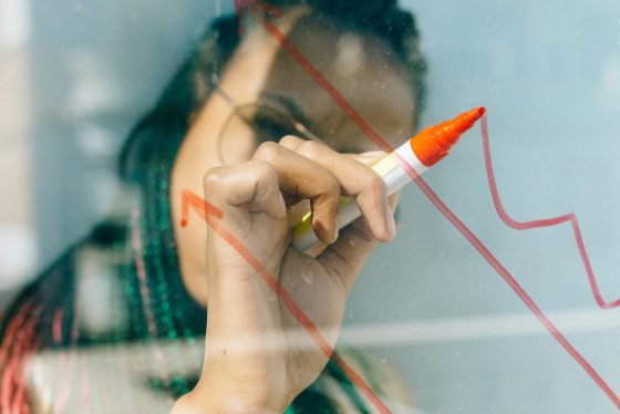 Person with braided hair drawing an upward trending line graph on a transparent surface with an orange marker,