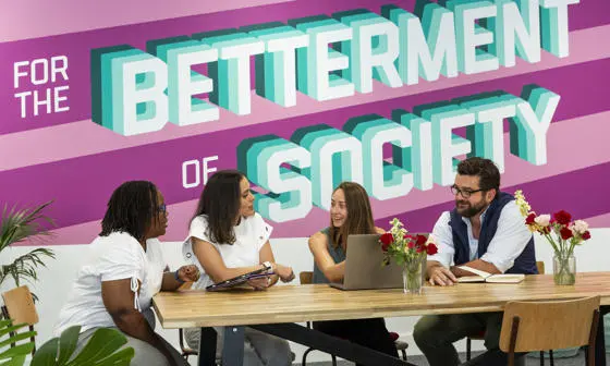 Four EMSBE students collaborating at a wooden table with laptop against a vibrant pink wall featuring "For the Betterment of Society" in bold 3D typography.