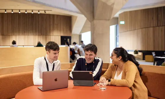Three EMSBE students collaborating with laptops at a round table outside lecture theatre.