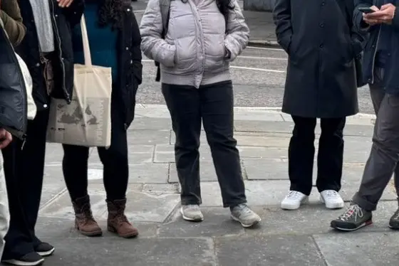 A group stand on a street corner as a woman gestures. Everyone wears warm coats, the sky is grey.