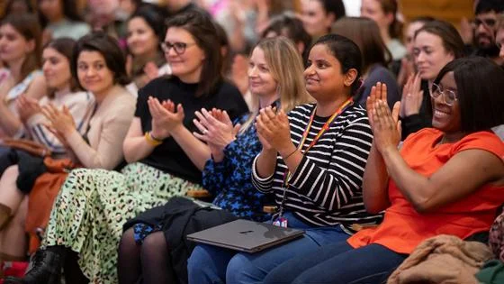The audience at an event clap and smile towards the stage.