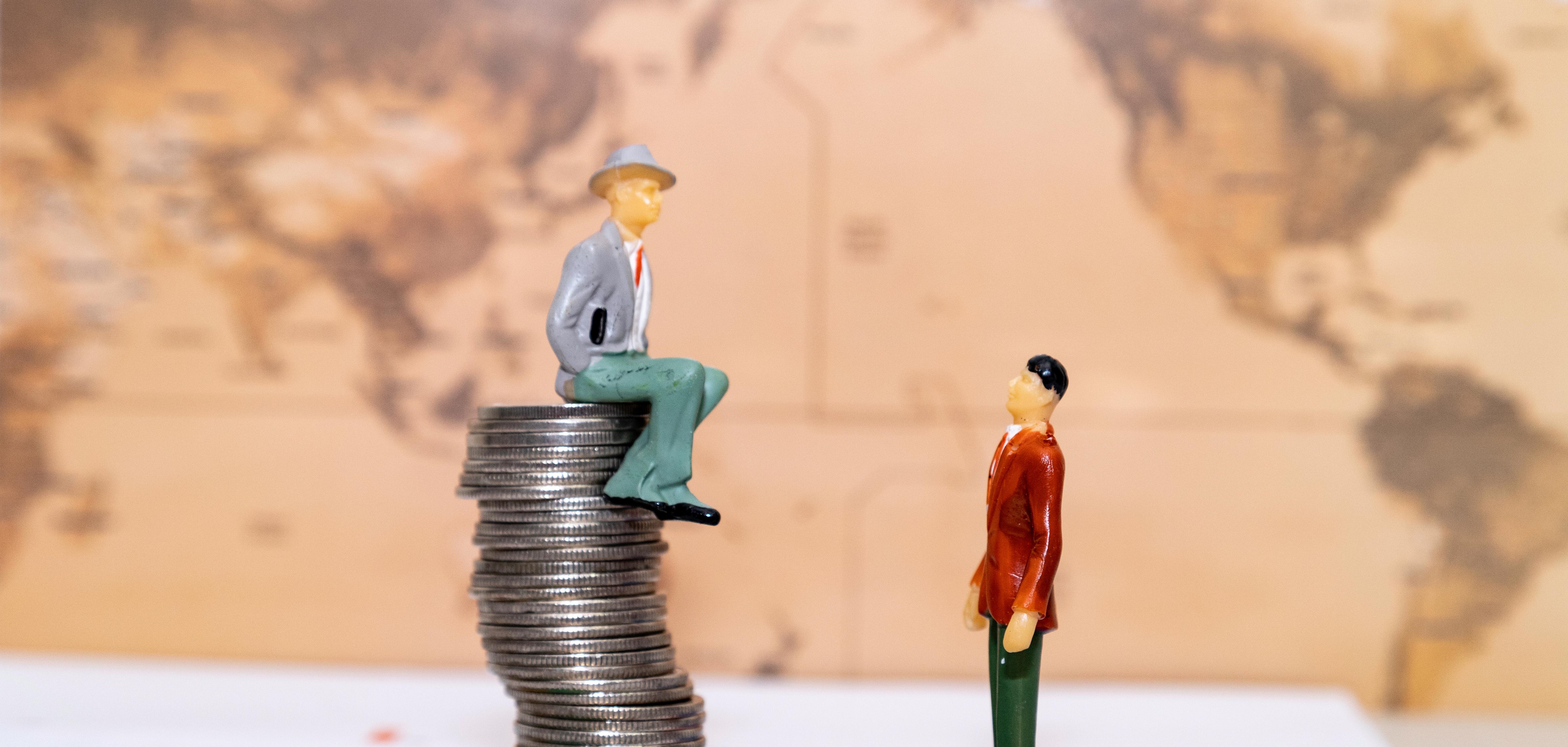 Model man sitting on a tower of coins looking down on another model man