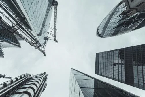 Upward view of modern glass skyscrapers and cranes in London’s financial district against a cloudy sky.