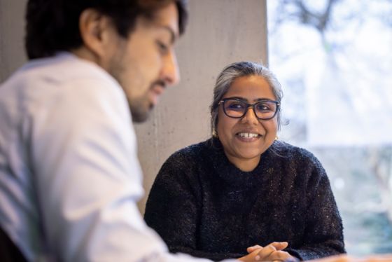 Two students sitting indoors, smiling and talking together in a quiet study area near a window.