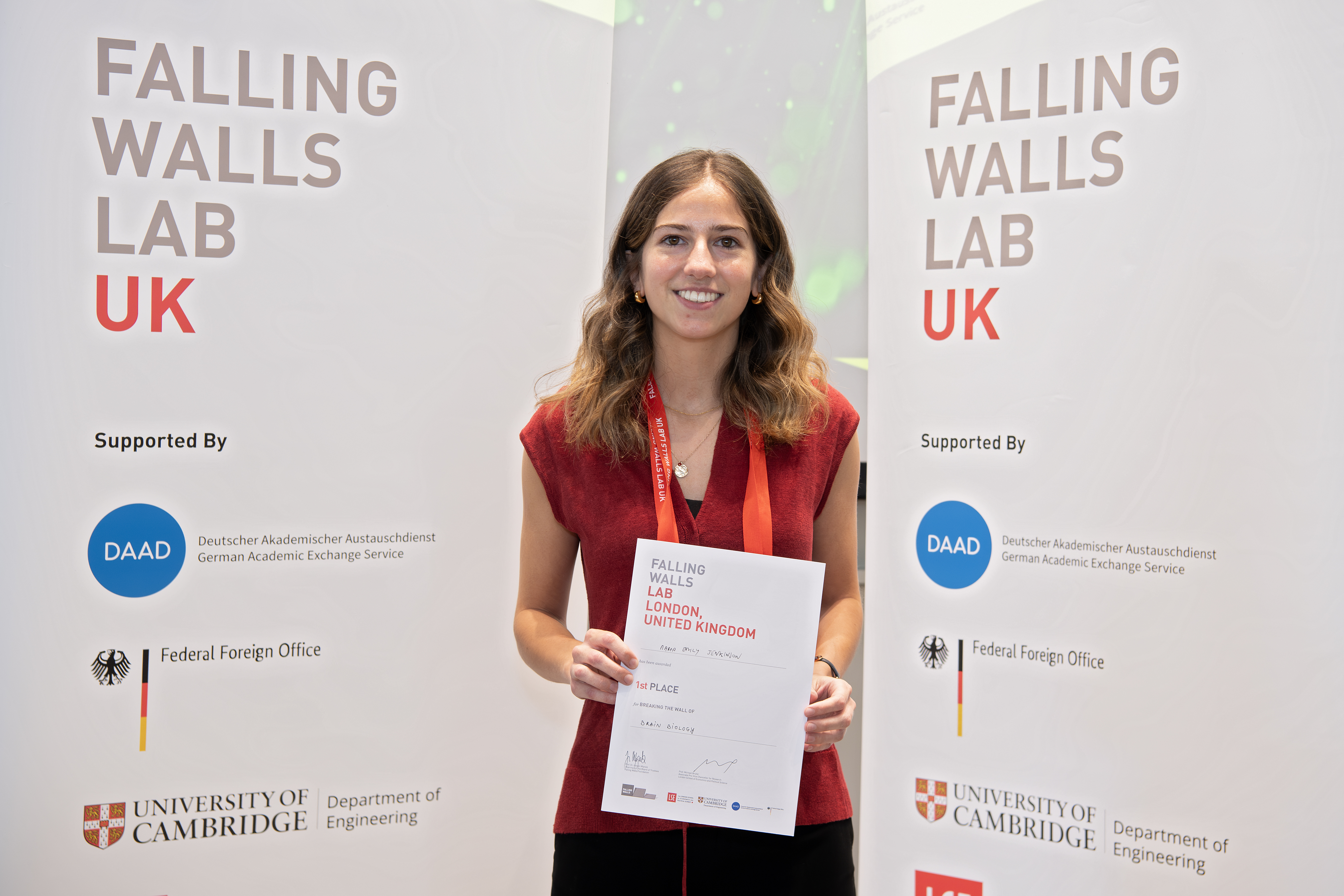 María Jenkinson Álvarez standing in front of the Falling Walls Lab UK sign, holding her 1st place certificate. 