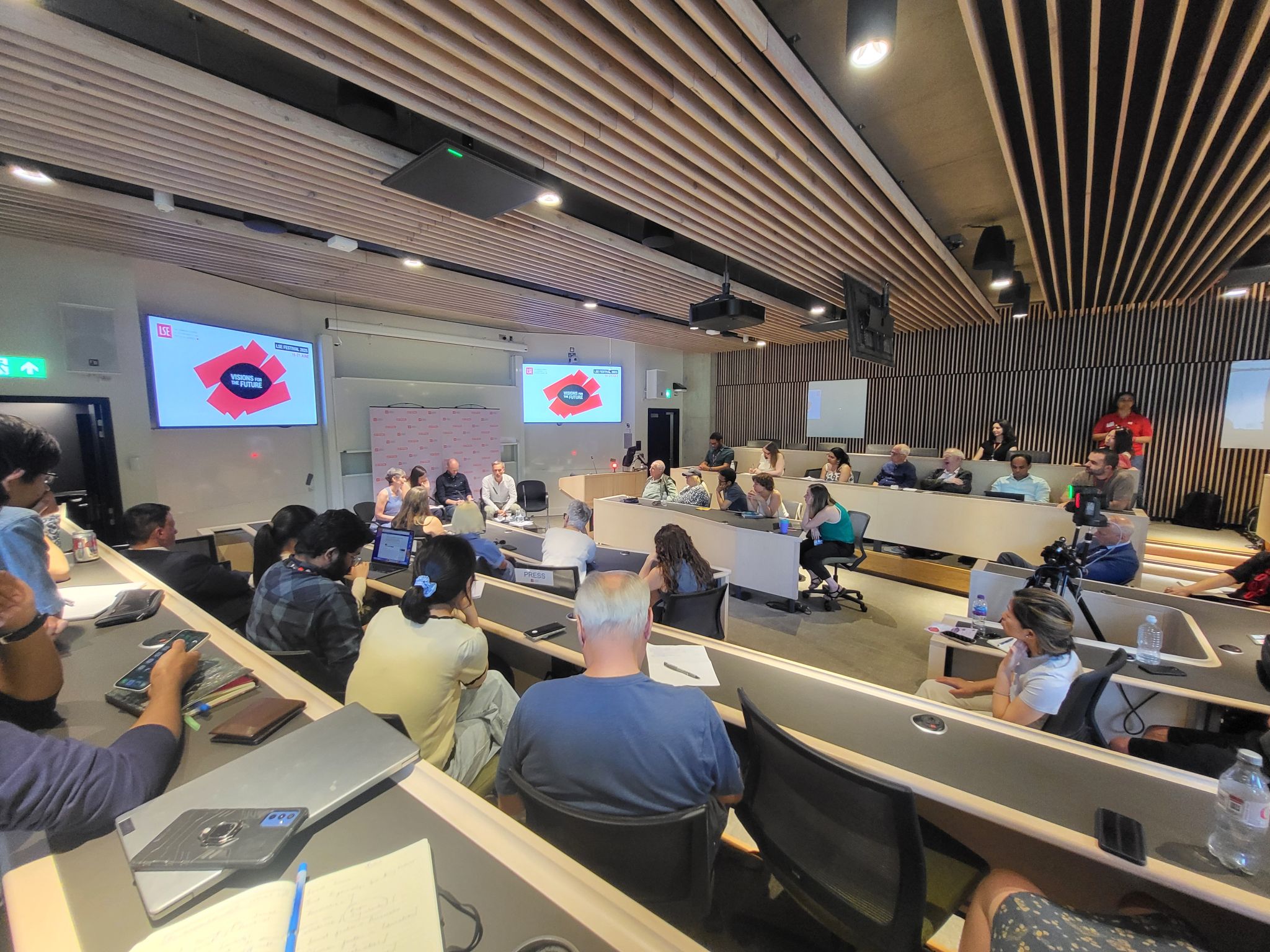 A large audience seated in a tiered lecture theatre during an LSE Festival event. Participants are gathered around a central discussion area while attendees watch from surrounding seats. Two presentation screens display the LSE logo and “Visions for the Future” branding.