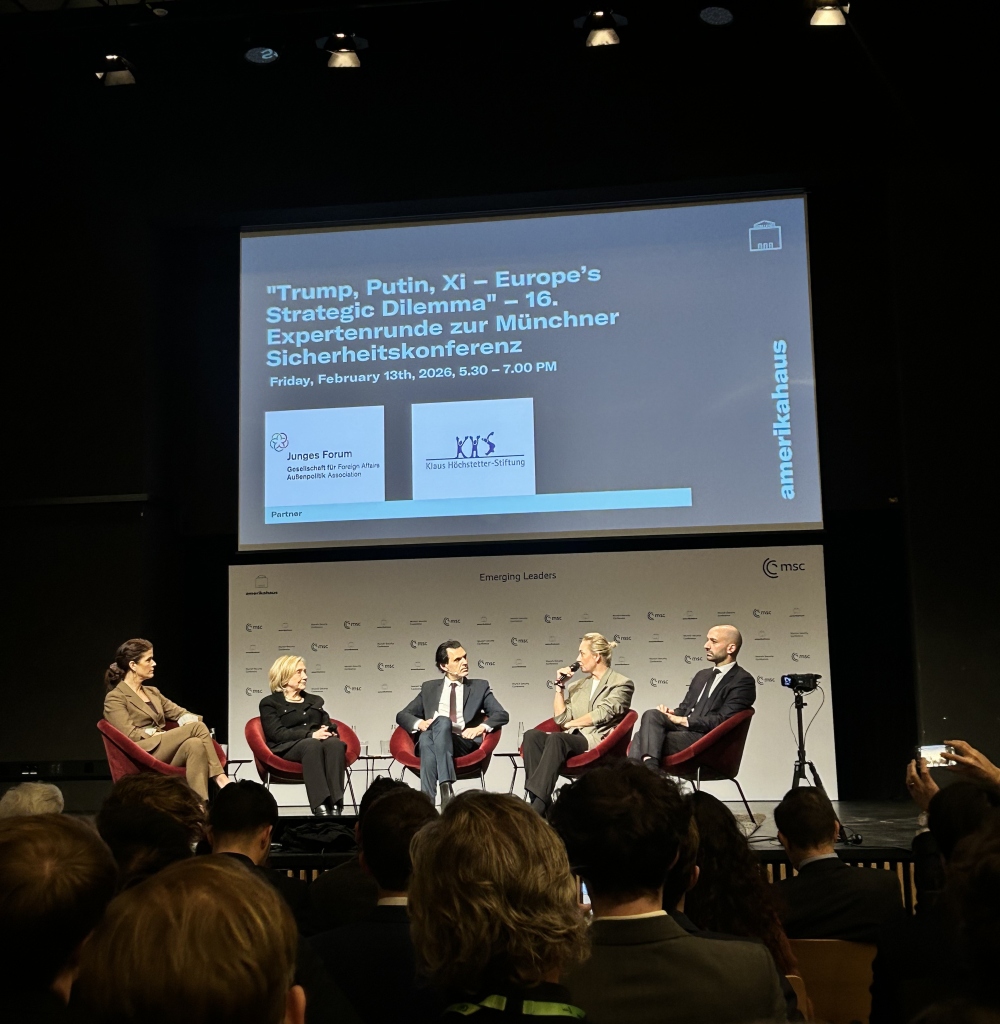 A panel of four speakers seated on stage in red chairs during a conference discussion, with a large projection screen above displaying information about a strategic security event. An audience is seated in the foreground, and conference branding is visible on the backdrop.