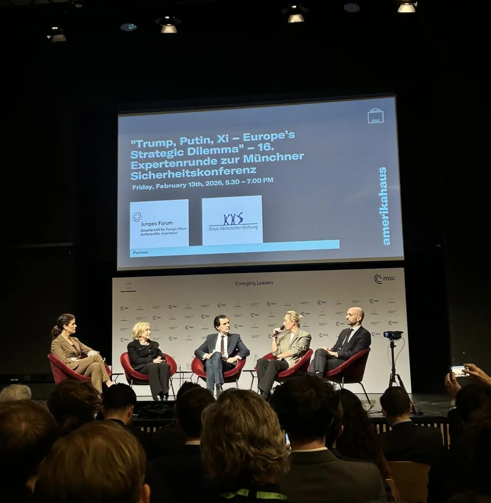 A panel of four speakers seated on stage in red chairs during a conference discussion, with a large projection screen above displaying information about a strategic security event. An audience is seated in the foreground, and conference branding is visible on the backdrop.