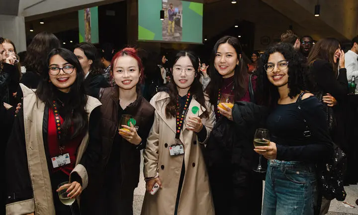 Five students at a Geography Welcome party holding drinks and smiling to camera