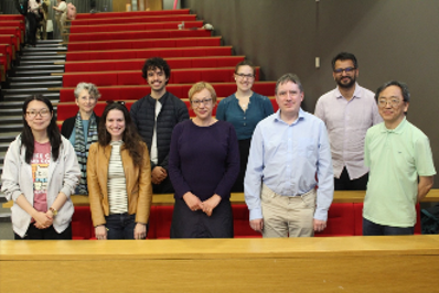 A group of nine people standing together in front of tiered red seating in a lecture theatre