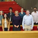 A group of nine people standing together in front of tiered red seating in a lecture theatre