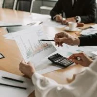 Professionals seated at a conference table reviewing printed financial charts using a calculator during a meeting.