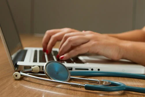 Stethoscope beside a person working on a laptop.