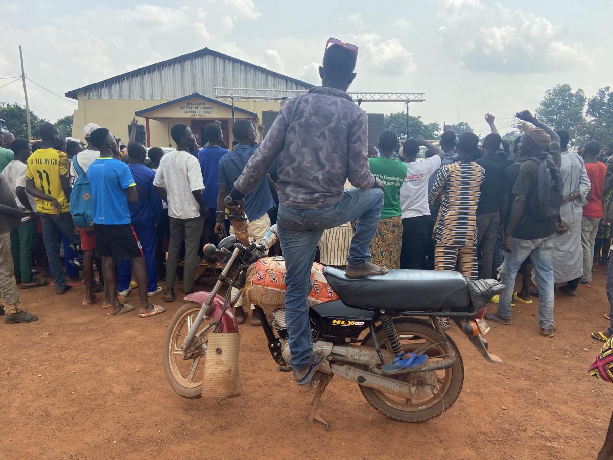 A group of people with their backs to the camera watching a screen. On person is standing on a motorbike to see above the crowd.